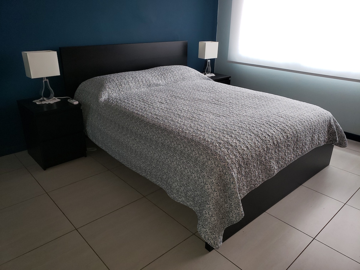 A spacious bedroom features a queen size bed with a light patterned coverlet. Two bedside tables hold modern lamps. Soft, natural light pours through a large window, illuminating the smooth tiled floor and creating a calm atmosphere.