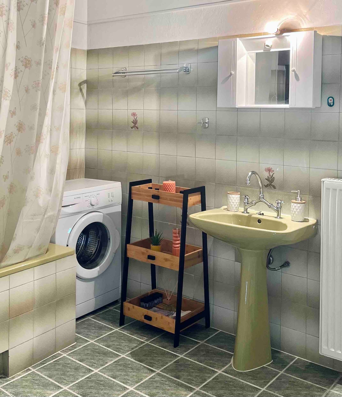The bathroom features a washing machine beside a modern sink with elegant fixtures. A small shelving unit holds neatly arranged items. Tile walls are complemented by floral accents, while soft lighting provides gentle illumination. A shower curtain adds privacy to the space.