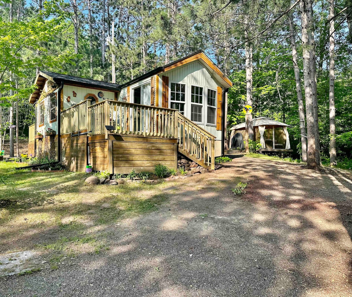 A cozy cabin is nestled among the trees, showcasing a wooden deck that leads to the entrance. Natural light filters through the surrounding greenery, and a screened gazebo is visible in the background, providing a serene outdoor space.