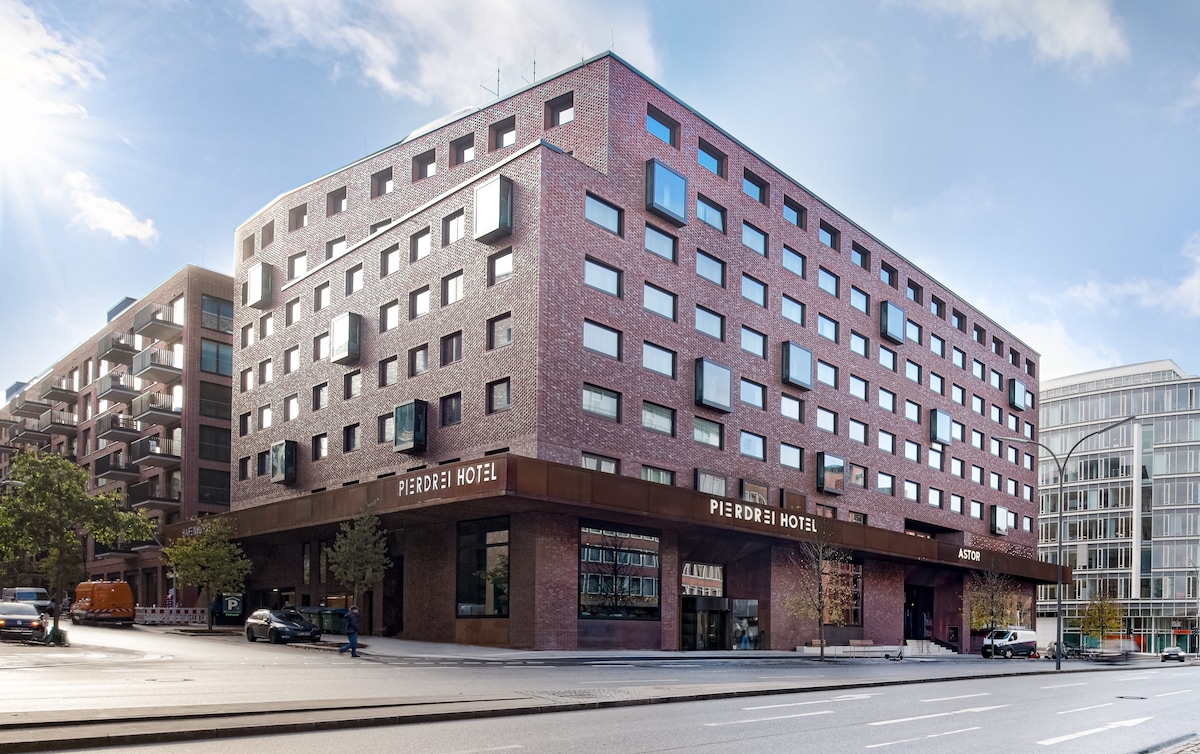 The exterior of PIERDREI HOTEL is characterized by a modern brick façade, featuring numerous large windows. The building’s rounded corners are complemented by a welcoming entrance marked with the hotel name. Surrounding trees and urban elements create a vibrant city atmosphere.