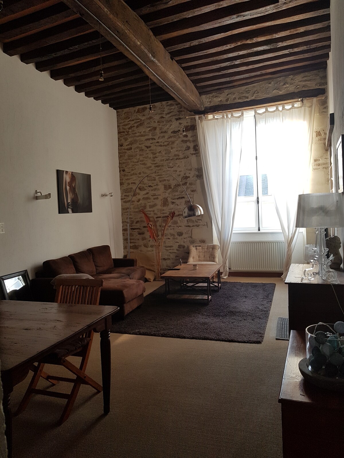 An inviting living area features exposed wooden beams and a stony wall, complemented by a brown sofa and a wooden coffee table. Natural light enters through a large window draped with sheer curtains, illuminating the space. A dining table with wooden chairs is visible to one side.
