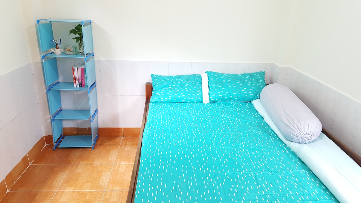 A bedroom is shown featuring a spacious bed covered with a bright blue sheet adorned with white patterns. A light blue pillow is positioned at one end of the bed. Adjacent to the bed, a minimalist shelving unit holds books and a small potted plant.