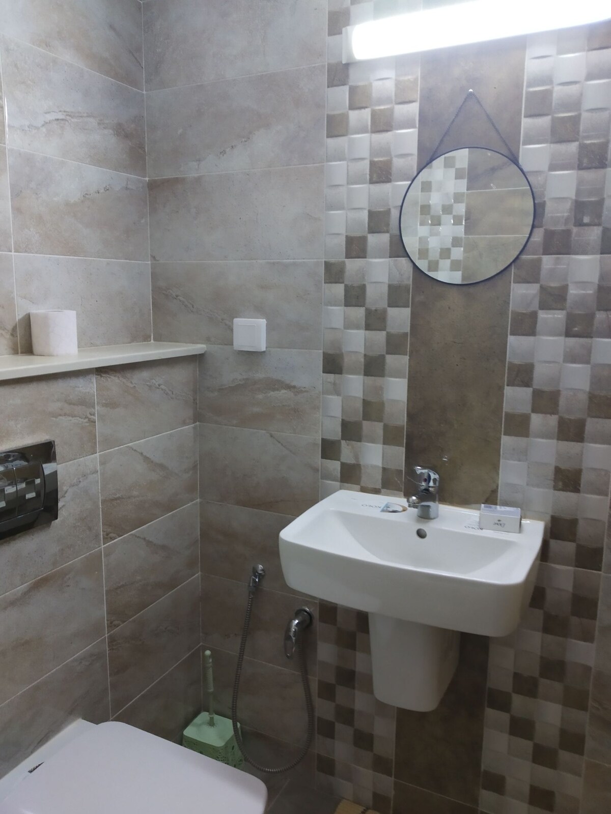 A bathroom featuring modern tiled walls with a neutral color scheme. A circular mirror is mounted above a compact sink, which is positioned beside a toilet. A small shelf is visible for toiletries, and soft lighting enhances the clean and fresh atmosphere.