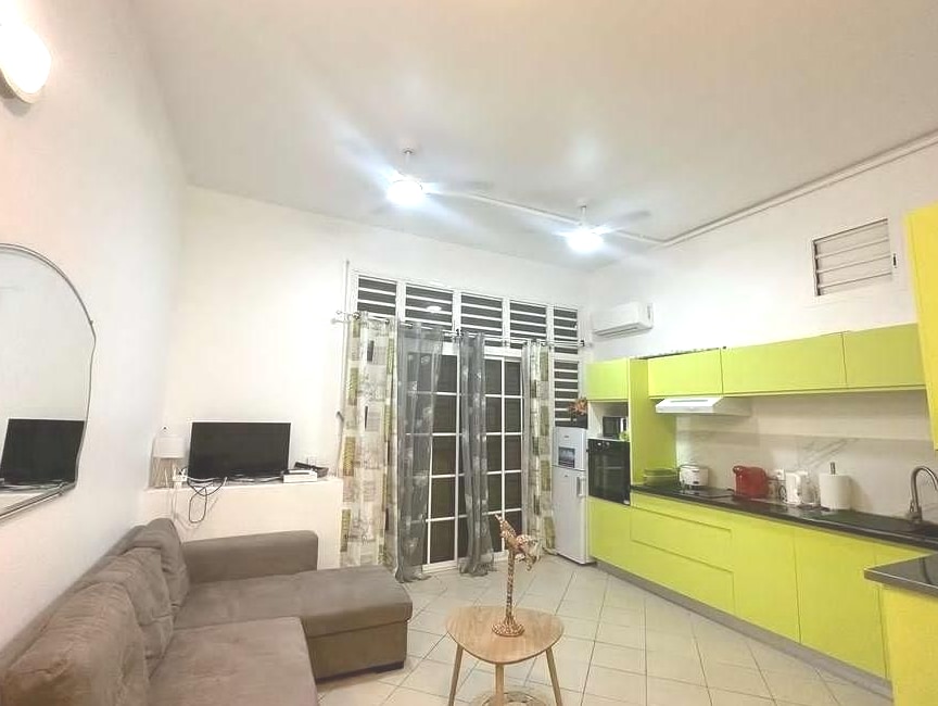 A modern kitchen space features bright green cabinetry and stainless steel appliances. A comfortable sectional sofa is arranged nearby, complementing the overall open layout. Natural light enters through large windows dressed with sheer curtains, illuminating the tiled floor.