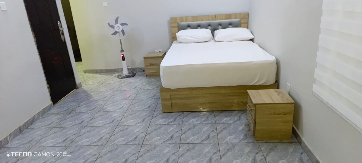 A well-furnished sleeping area features a spacious bed with white linens, complemented by two wooden nightstands. A standing fan is positioned next to the bed, and the room is illuminated by natural light filtering through a partially covered window.