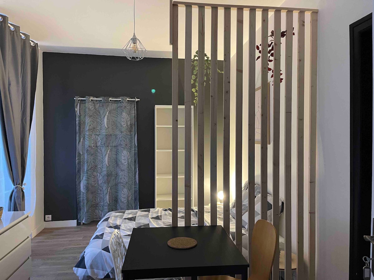 A divided space is featured, showcasing a cozy sleeping area behind a wooden slatted partition. A double bed is covered with a patterned comforter, while natural light enters through the curtains. A small black table with wooden chairs is positioned in the foreground.