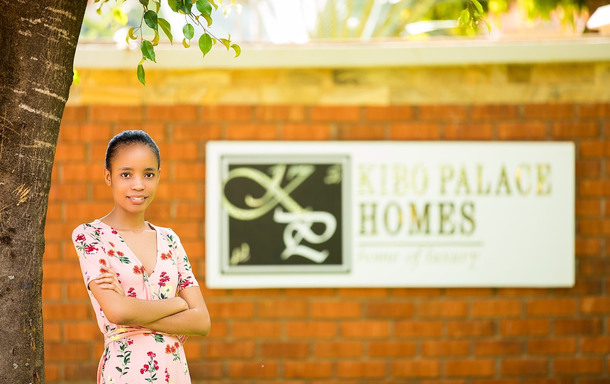 A welcoming entrance to Kibo Palace Homes is displayed, featuring a well-maintained brick wall. The name of the property is presented on a prominent sign, with lush greenery partially framing the scene, enhancing the inviting exterior.