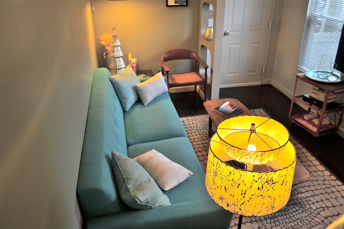 A modern living area features a soft blue couch accented with colorful pillows. A wooden chair and a coffee table with books are present. A unique yellow lamp provides warm lighting, and a door leads to the outdoors, with soft natural light filtering through nearby windows.
