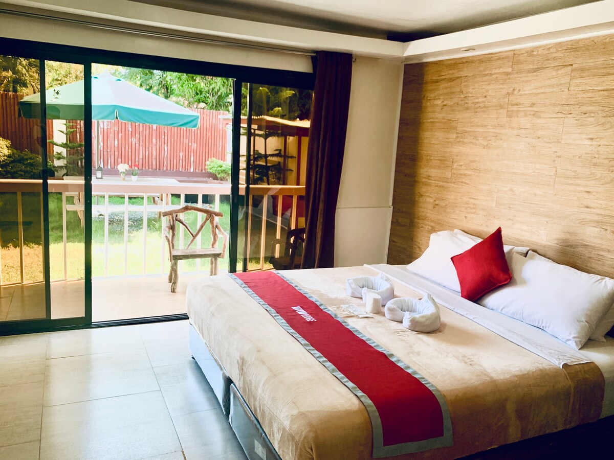 A spacious bedroom is highlighted by a large bed with crisp white linens and a decorative red accent pillow. Floor-to-ceiling sliding doors open to a garden view, allowing natural light to fill the room. A wooden chair is visible outside on the patio.