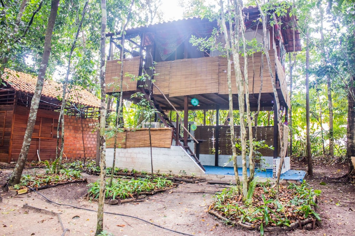 A two-story wooden structure is surrounded by lush greenery, showcasing a tranquil setting. The ground level features an open area with blue flooring, while wooden decking is visible on the upper level. Nearby, a landscaped area with low vegetation adds to the serene environment.