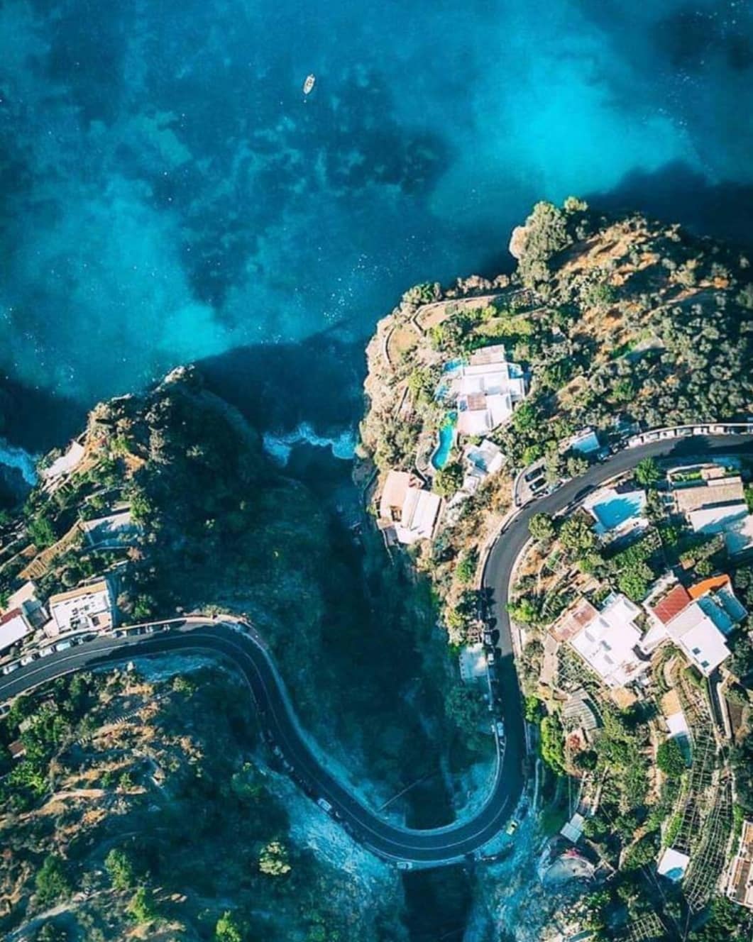 An aerial view captures the winding road adjacent to the coastline, surrounded by lush greenery and colorful homes. The clear blue waters contrast with the landscape, highlighting the tranquil natural beauty of the area. The scene emphasizes the proximity to both land and sea.