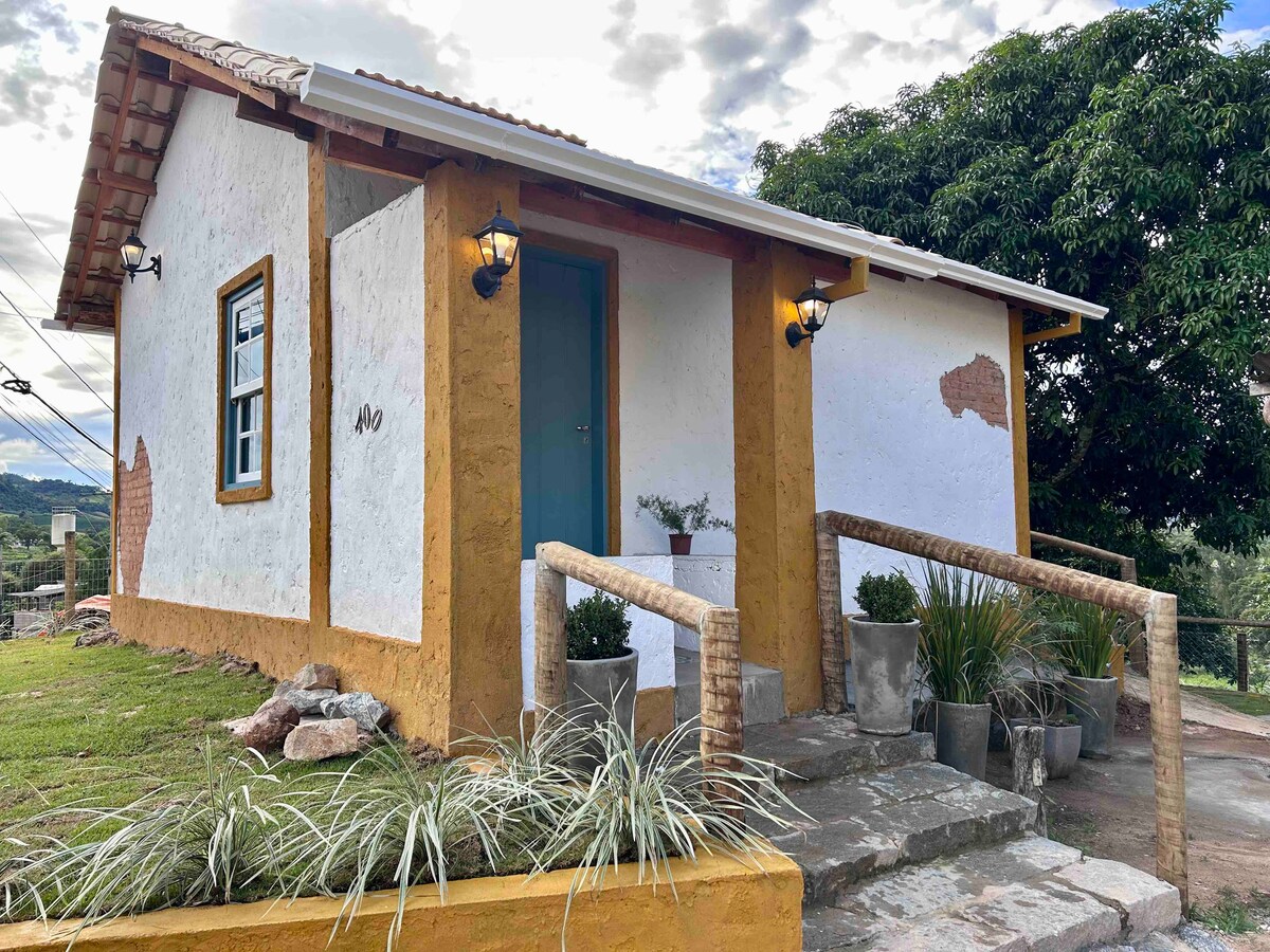 A charming exterior of a renovated 1950s house features a yellow and white color scheme. The entrance is marked by a blue door and two lanterns. Plants are placed in decorative pots by the door, surrounded by a well-maintained grassy area.