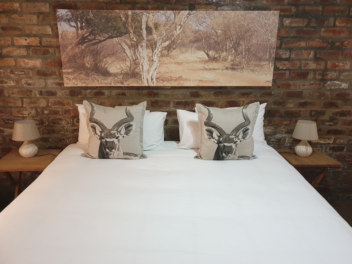 A spacious bed is adorned with neutral-toned linens and decorative pillows featuring wildlife imagery. Two bedside lamps with soft lighting complement the rustic charm created by the exposed brick wall and a photographic artwork depicting a serene landscape above the bed.