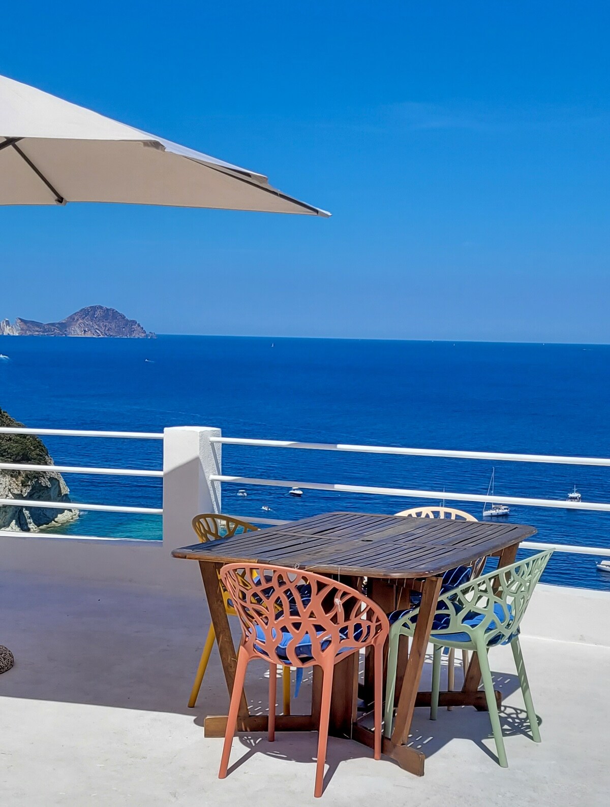 An outdoor dining area features a wooden table surrounded by colorful chairs in shades of pink, green, and beige. A large umbrella provides shade, with a clear view of the blue sea and distant boats on the water.