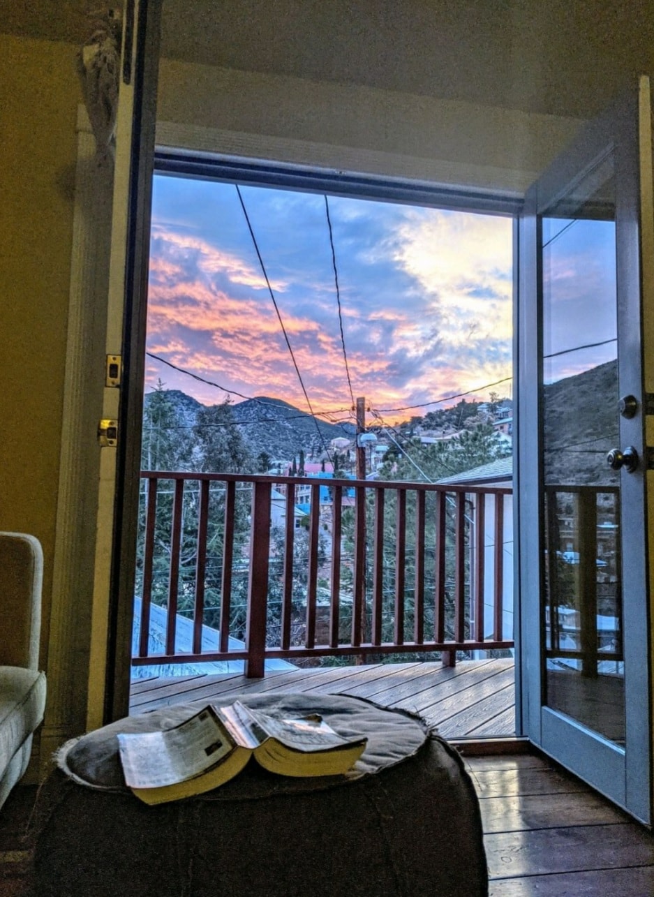 A view is captured through open double doors, revealing a vibrant sunset over the mountains. Soft pink and orange hues fill the sky. A cozy ottoman rests in the interior space, holding a few books, while the wooden deck is visible beyond.
