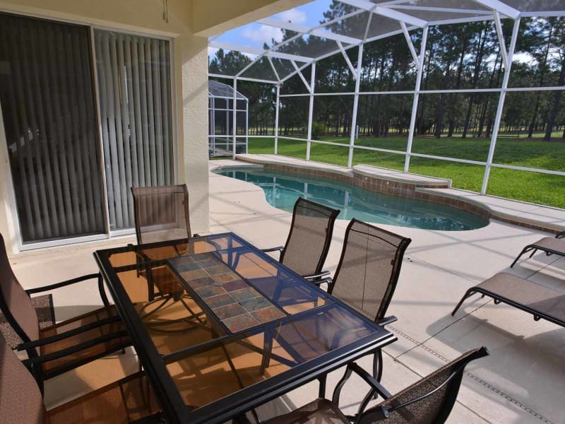 An outdoor patio area features a dining table surrounded by six chairs, with a glass tabletop that highlights a decorative design. A private pool is visible nearby, enclosed by a screened lanai, with a lush green lawn and trees providing a peaceful backdrop.