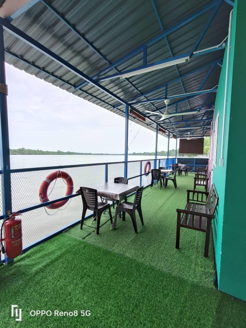 An outdoor seating area is displayed, featuring several tables and black chairs arranged along a walkway beside the river. A bright green carpet covers the floor. Two life rings and a fire extinguisher are visible, providing safety features in the environment.