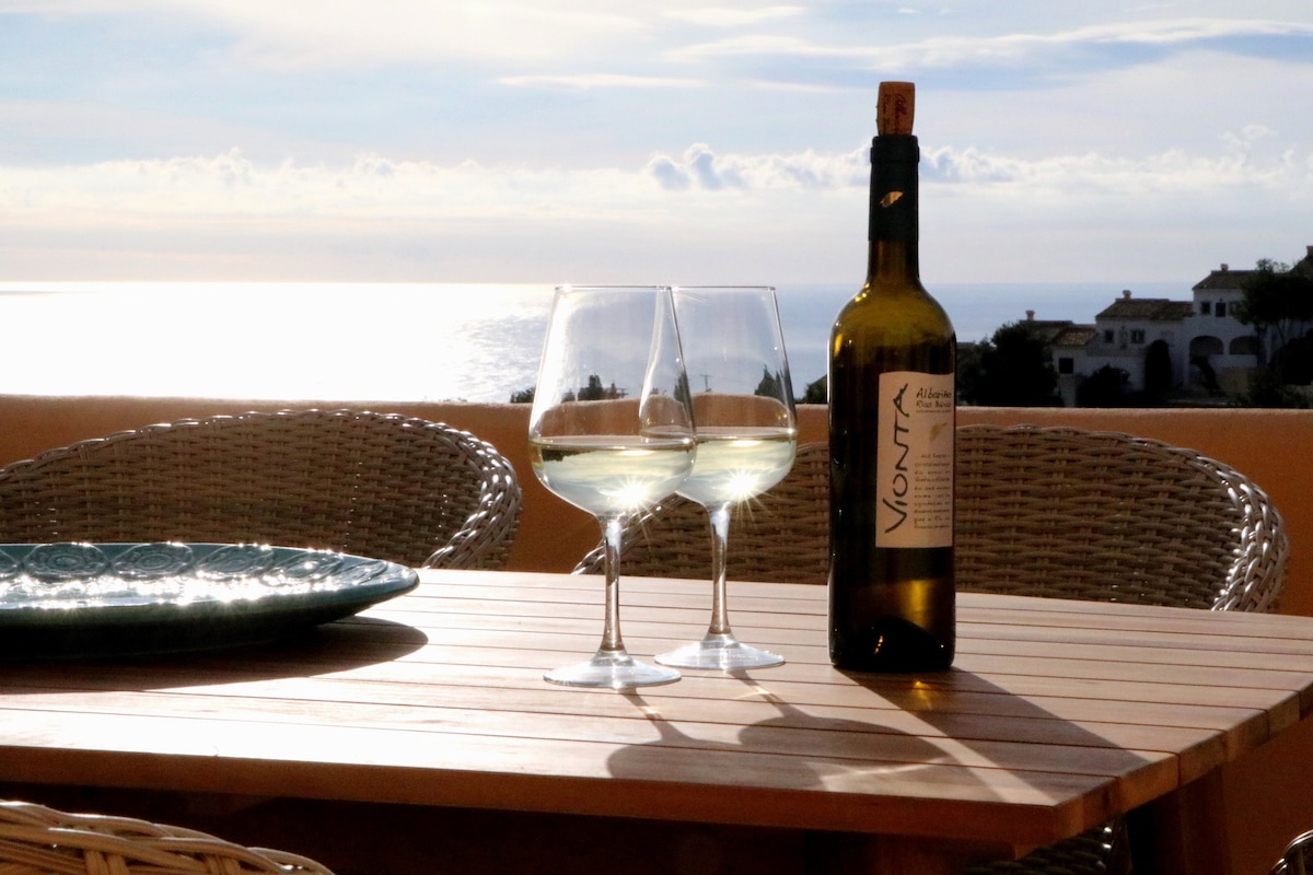 A wooden table is set for relaxation, featuring two wine glasses filled with white wine and a bottle of Vionta. A decorative plate sits nearby, with a backdrop of a shimmering sea reflecting soft light under a cloudy sky.