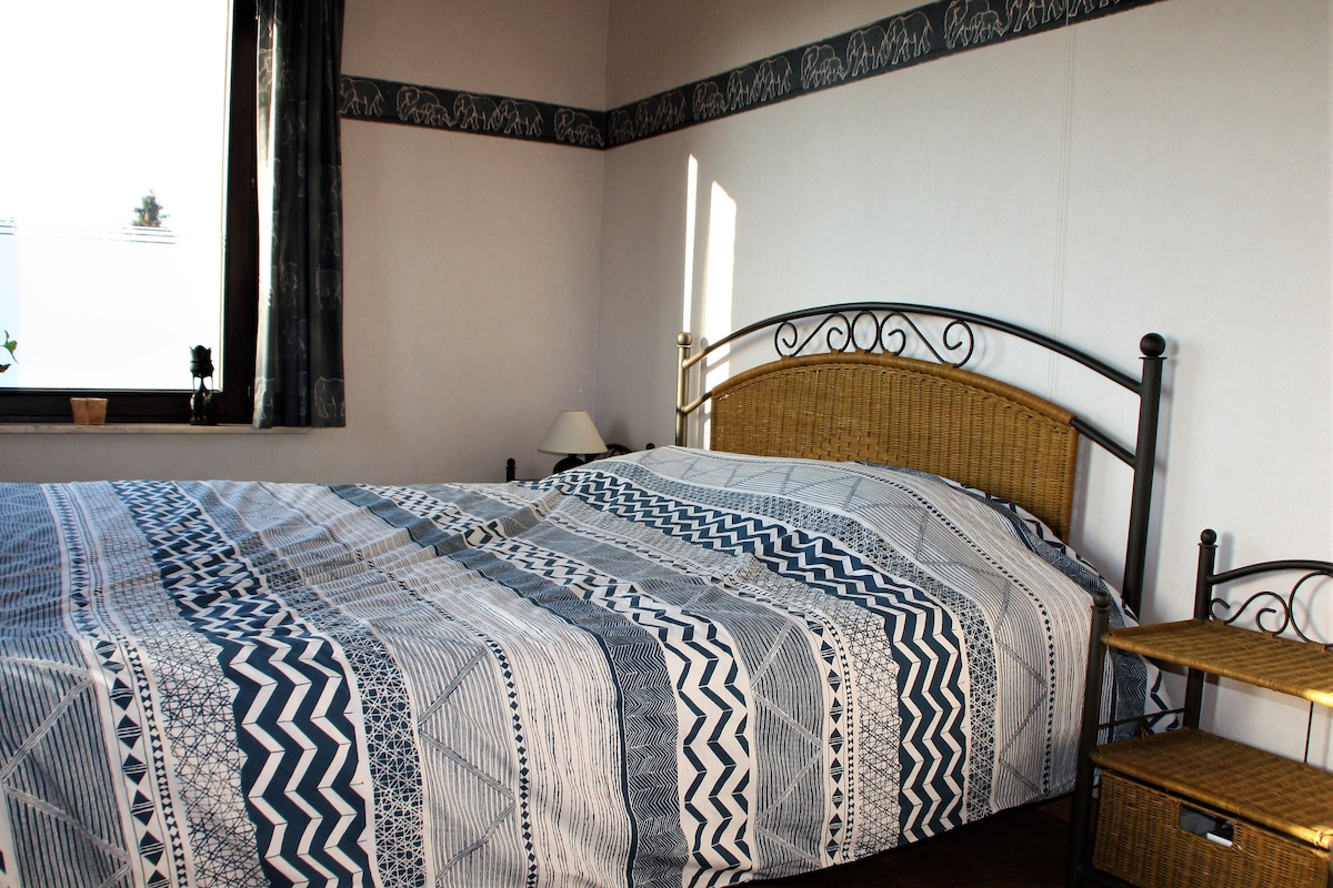The room features a spacious bed with a patterned duvet cover, complemented by two nightstands. Natural light enters through the window, and a small lamp rests on one nightstand, adding to the room's functionality and comfort.