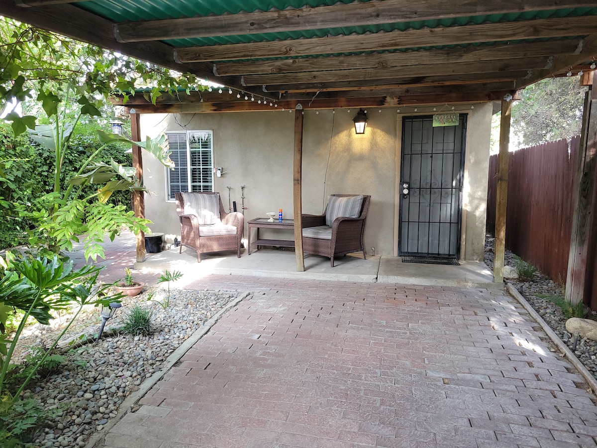 A shaded outdoor seating area is presented, featuring two wicker chairs and a small table. The space is adorned with various plants and surrounded by decorative stones. A solid door and window are visible, providing access to the studio.