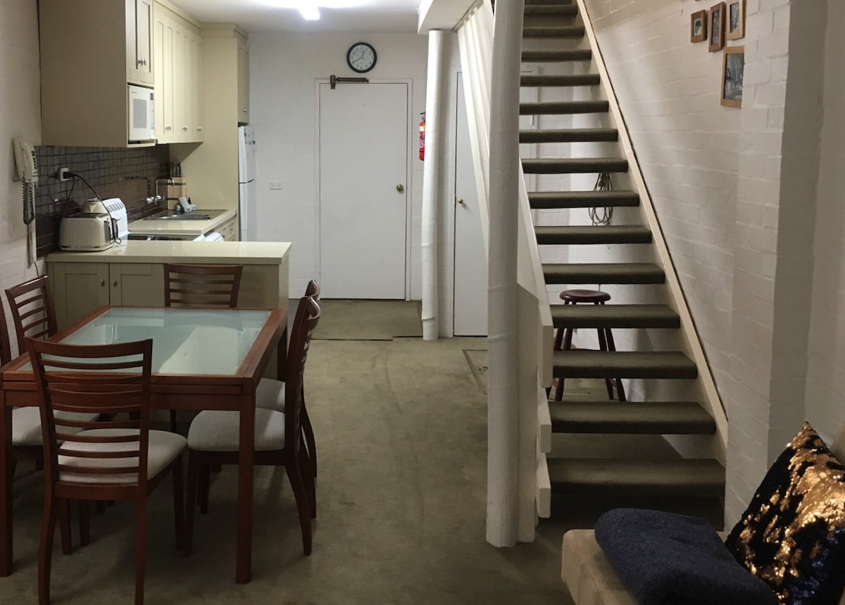 The dining area features a wooden table with six chairs beside a modern kitchen equipped with a refrigerator and microwave. A staircase leads to the upper level, while a clock hangs on the wall, and a door provides access to the rest of the apartment.