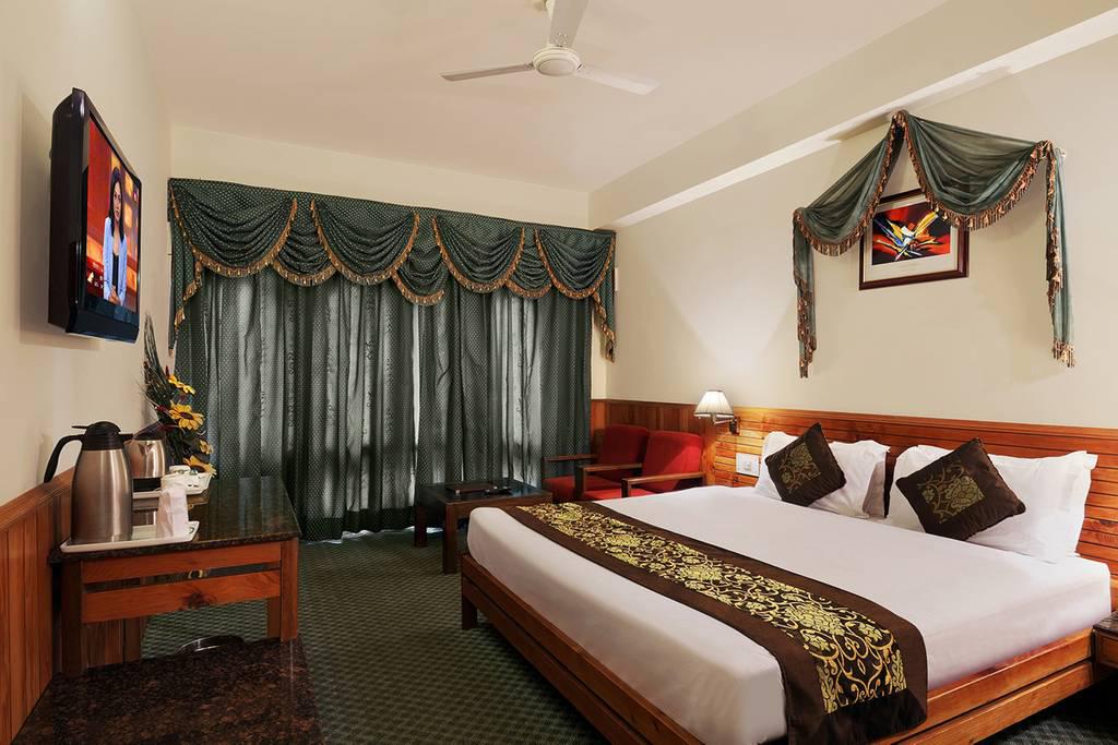 A comfortable room features a spacious bed with decorative cushions, a dark wooden headboard, and a TV mounted on the wall. Plush curtains frame a window, allowing soft light to filter in. A seating area includes a chair, table, and a kettle on a sideboard.
