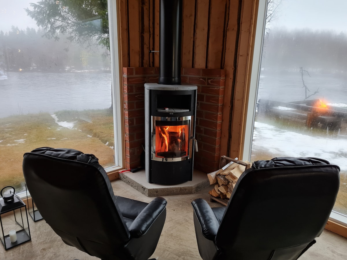 Two black chairs are positioned near a wood-burning stove, with flames visible through the glass front. The surrounding windows offer views of the river, shrouded in a misty atmosphere. Wood is neatly stacked beside the stove, contributing to a cozy ambiance.
