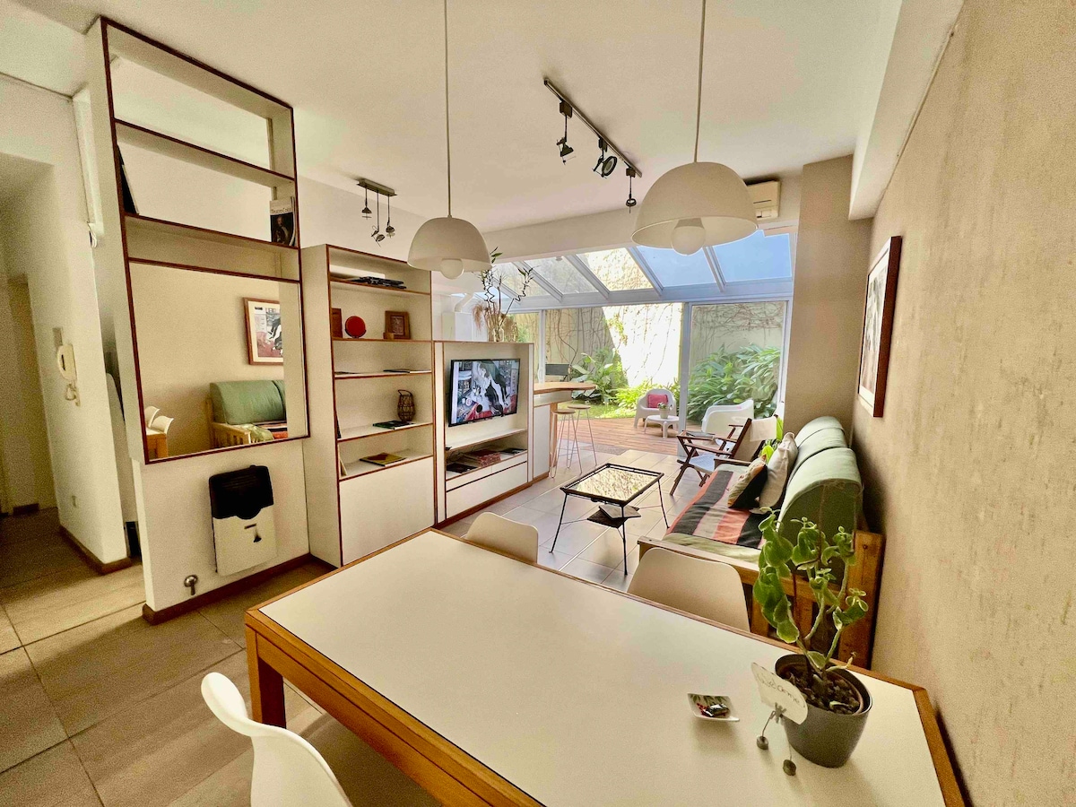 A bright living space features a white dining table with chairs, complemented by a cozy seating area that overlooks a lush garden. Large windows allow natural light to fill the room, while a television is mounted on a minimalist shelving unit.