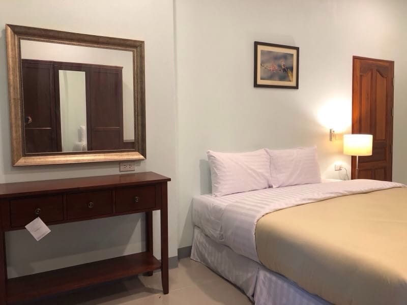 A cozy bedroom features a spacious bed with light linens, complemented by a warm blanket. A wooden console table is positioned against the wall, with a large mirror above it reflecting the softly lit space. A framed artwork and wooden door enhance the inviting atmosphere.