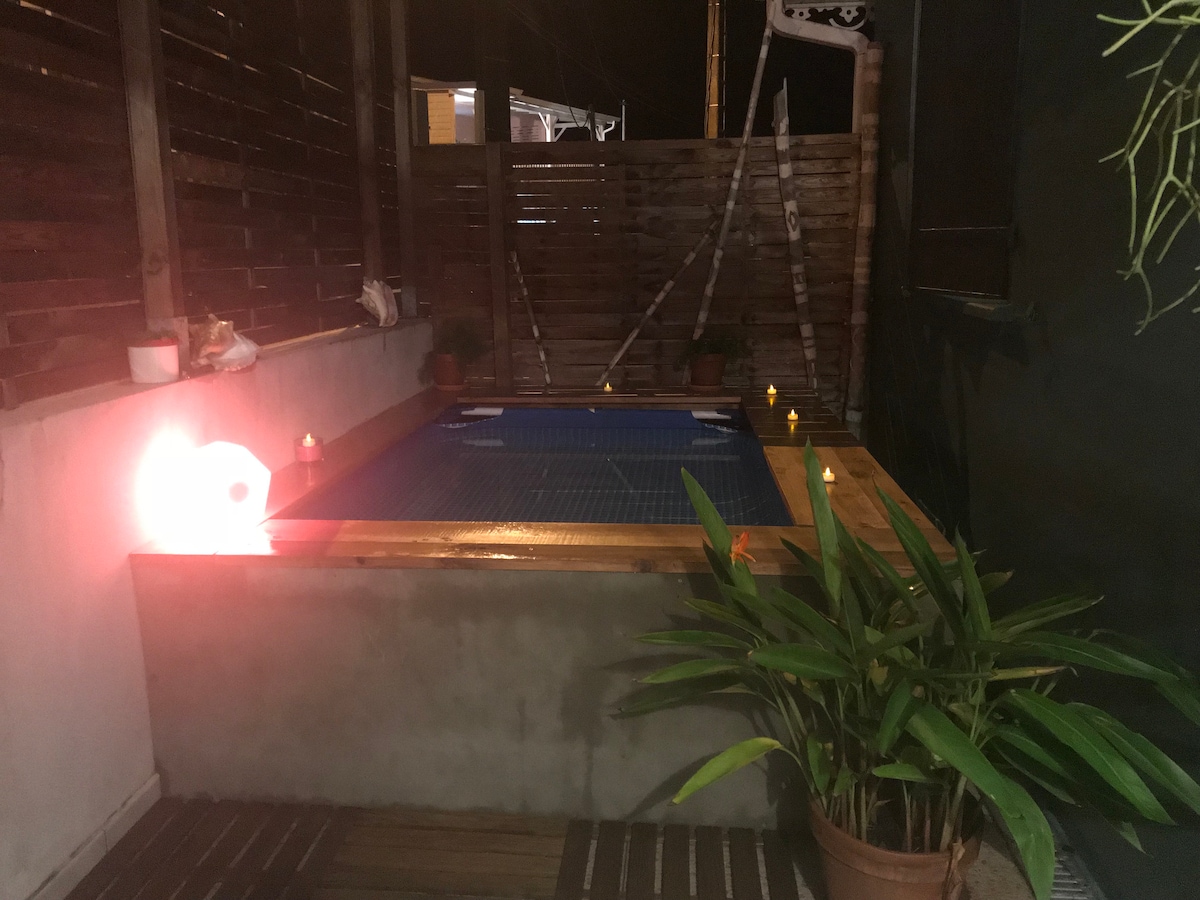 A private terrace features a small plunge pool surrounded by wooden decking. Potted plants are placed nearby, and the inviting atmosphere is enhanced by small candles lining the edge of the pool, suggesting a tranquil evening setting.