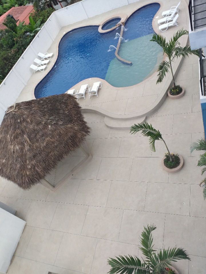 An aerial view of a pool area is presented, featuring a curvy swimming pool with a connected shallow section. Surrounding the pool are several lounge chairs positioned on light-colored tiles. A thatched-roof gazebo adds a shaded spot, accompanied by potted plants along the edges.