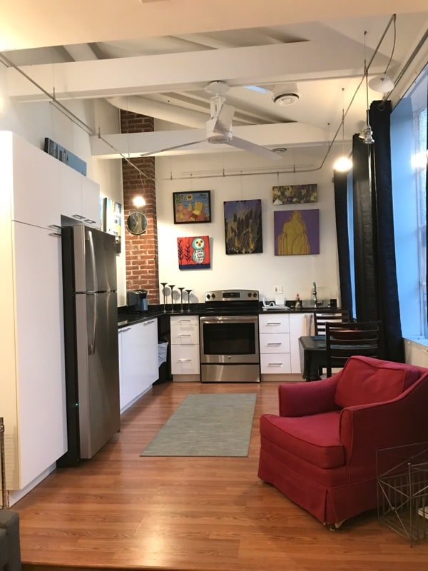 A functional kitchen area features modern appliances, including a refrigerator and stove, against a backdrop of exposed brick walls. Colorful artwork adorns the walls, while a cozy red chair adds a welcoming touch to the space. Large windows allow ample natural light to fill the room.