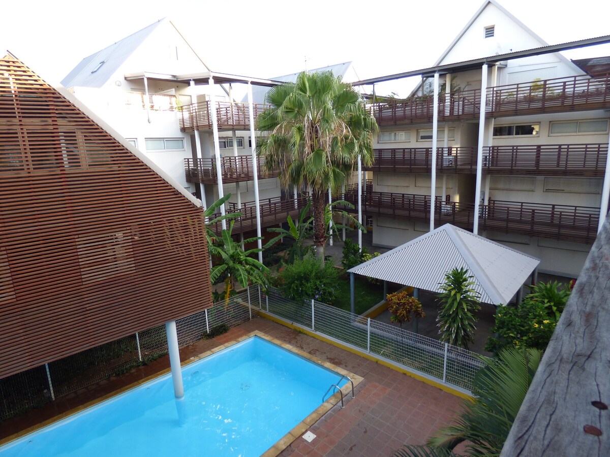 The image captures a view of the swimming pool surrounded by tropical landscaping in the courtyard. Three levels of the building are visible, with wooden balconies and palm trees providing shade. A pergola is seen near the pool, offering a space for relaxation.