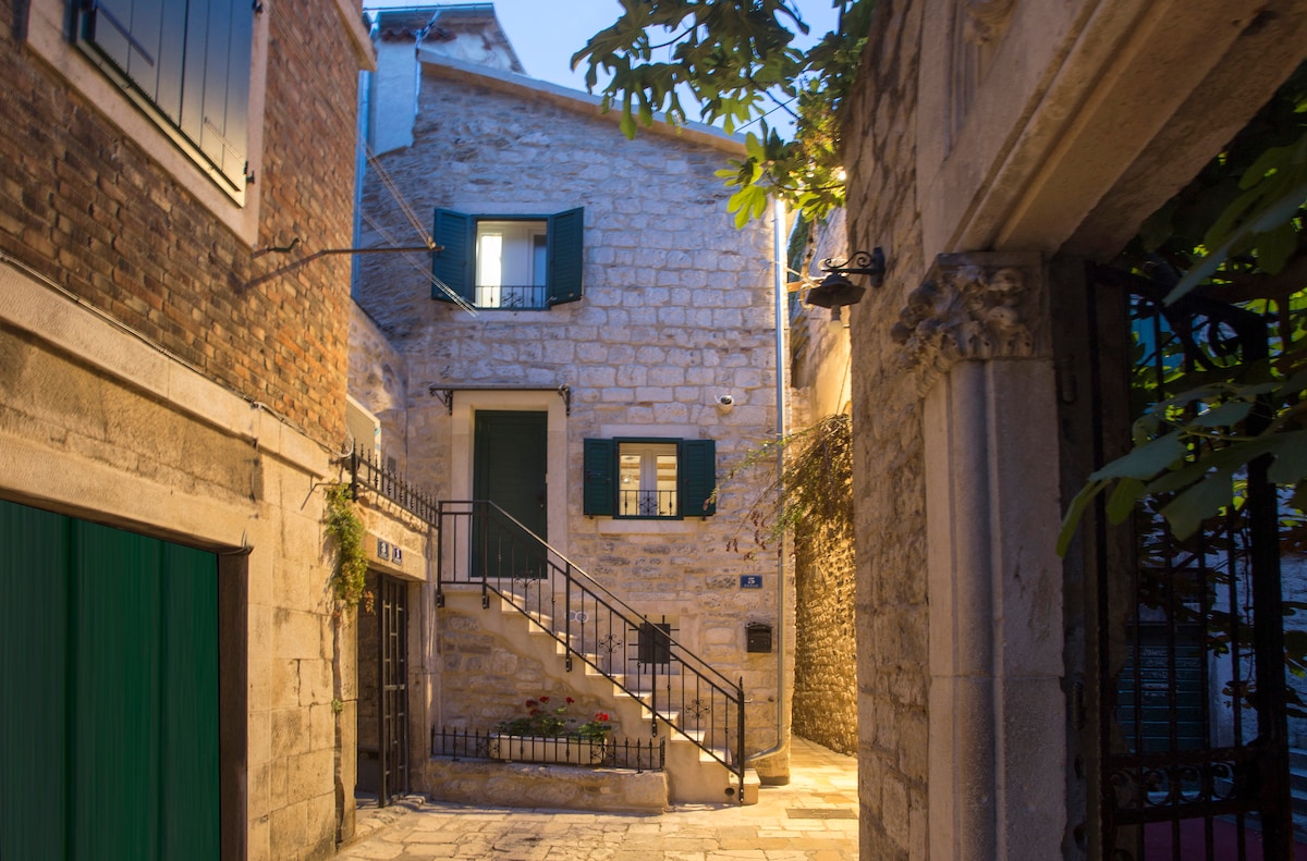 A quaint stone building is flanked by narrow cobblestone pathways and greenery. Green shutters adorn the windows, while an iron staircase leads to the upper level. Soft lighting enhances the architectural features, creating a calm and inviting street scene.