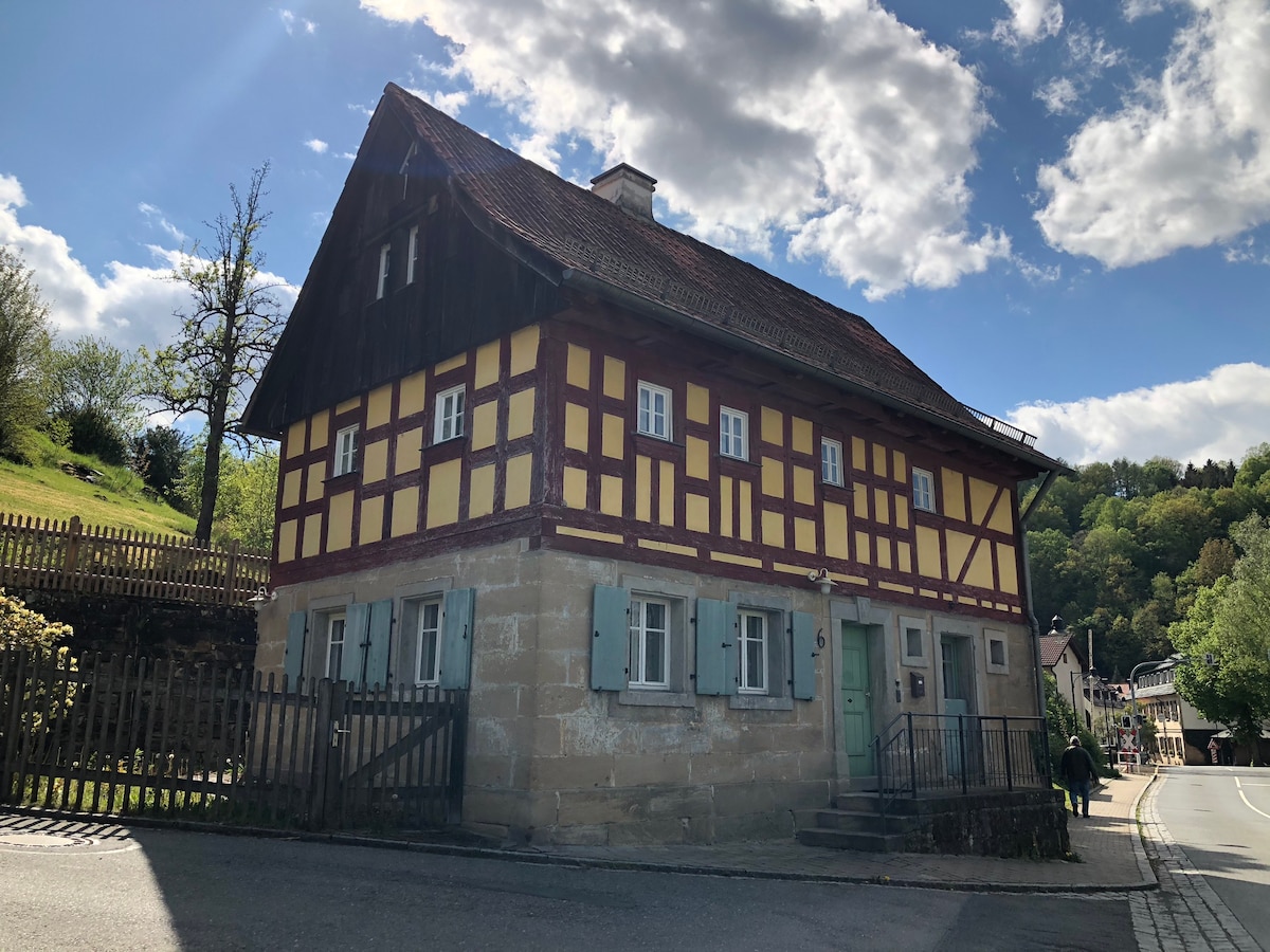The charming house features a combination of half-timbered construction and a stone base, showcasing a yellow facade with wooden accents. The peaked roof and multiple windows add to its character. A wooden fence surrounds the property, providing a quaint and inviting entrance.