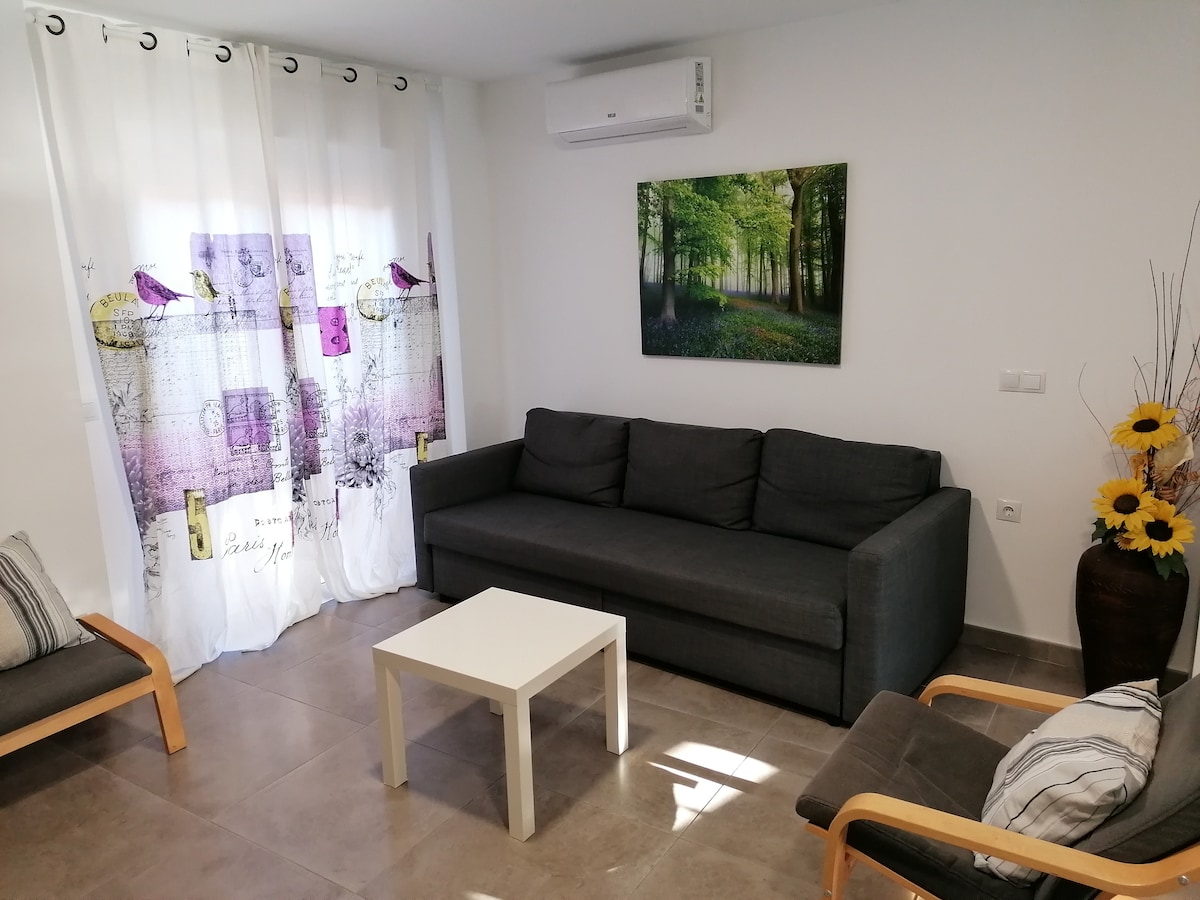 A bright living area features a comfortable gray sofa and two chairs beside a white coffee table. Light flows in through sheer curtains adorned with floral motifs, enhancing the space's openness. A vibrant nature-themed artwork is displayed on the wall.