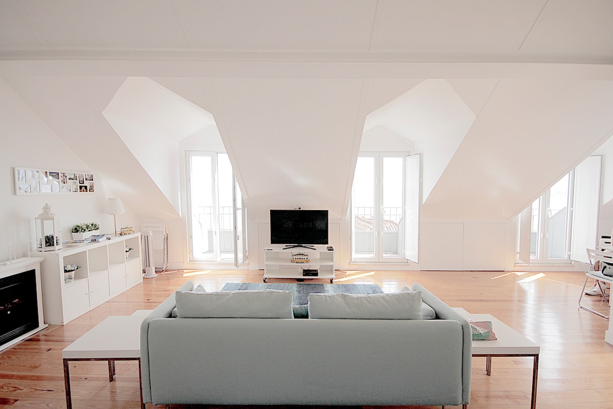 An open living space features a light-coloured sofa facing a television. Large windows allow natural light to fill the room, creating an airy ambiance. The walls are white, and the wooden floor adds warmth to the environment. Shelving units provide opportunities for decorative touches.
