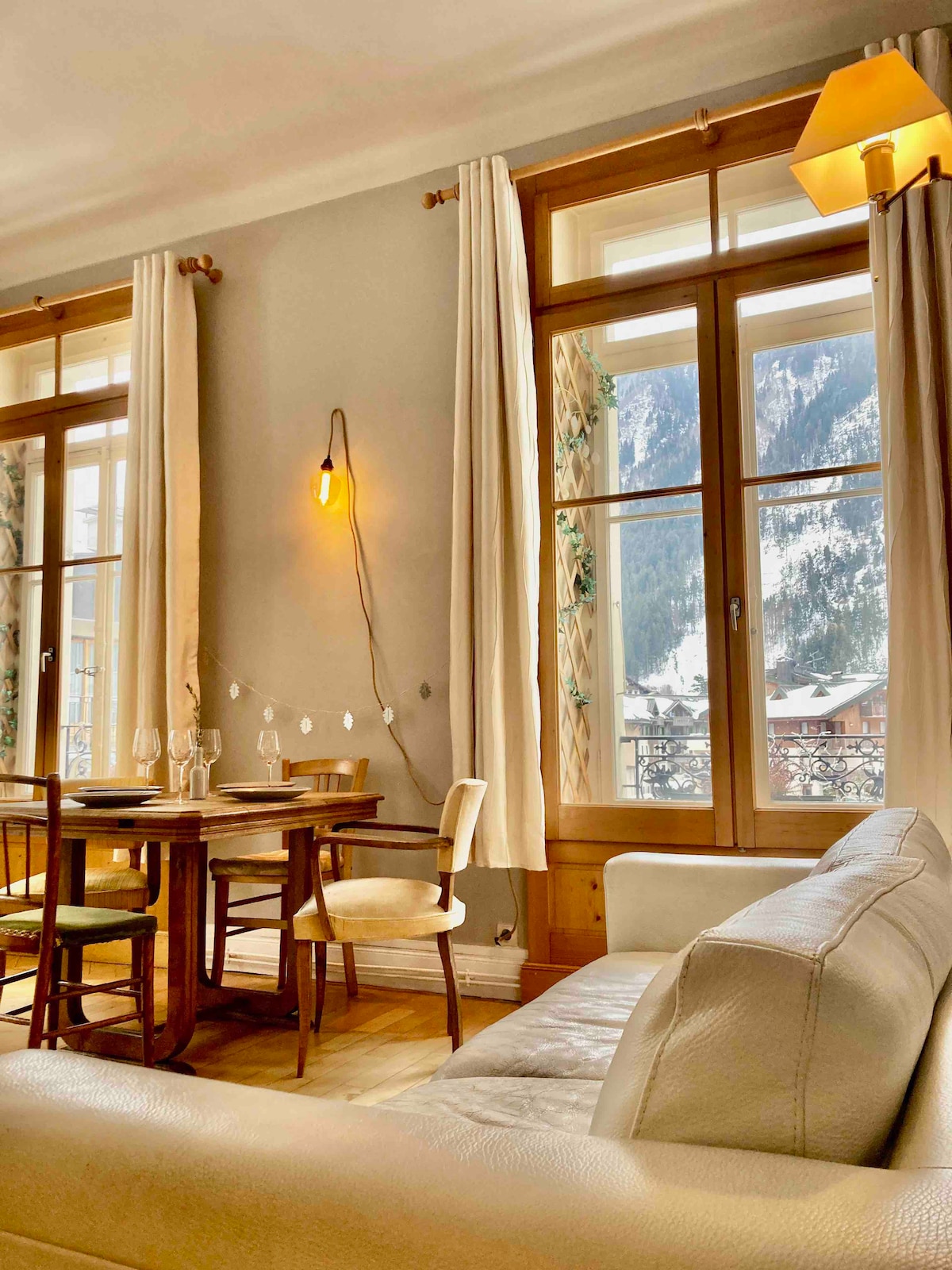 The open living and dining area features large windows that frame scenic mountain views. A wooden dining table is surrounded by chairs, with soft lighting from a wall lamp and nearby floor lamp enhancing the space. Natural light fills the room, creating a warm atmosphere.