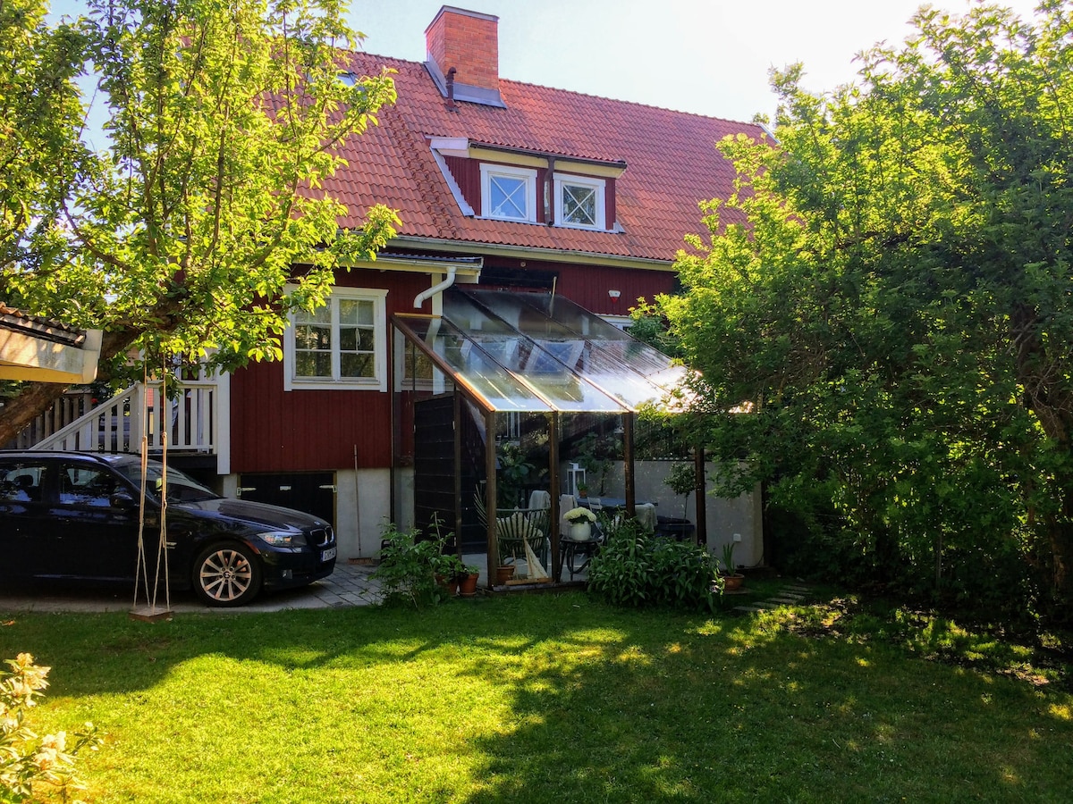 A red wooden semi-detached house features a glass orangeri surrounded by greenery. A private garden space is well-maintained, with ample grass and trees providing shade. A black car is parked nearby, completing the serene outdoor setting.