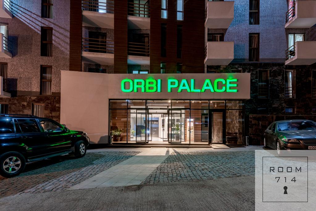 The entrance of the Orbi Palace is illuminated with bright green signage. Large glass doors welcome guests, flanked by sleek black vehicles parked in front. The modern facade displays a mix of textures, with stone and concrete elements contributing to its contemporary appearance.