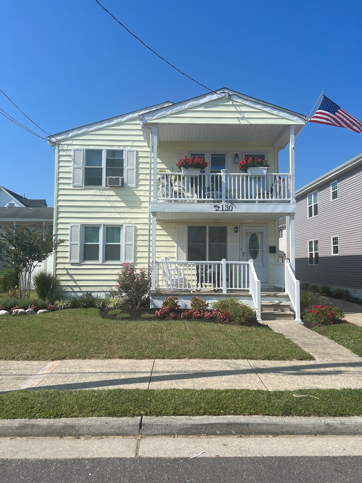 A two-story home is displayed, featuring light yellow siding and white railings. An American flag waves above the front porch, where potted plants add color. Lush green grass surrounds the walkway leading to the entrance, complemented by flower beds along the edge.