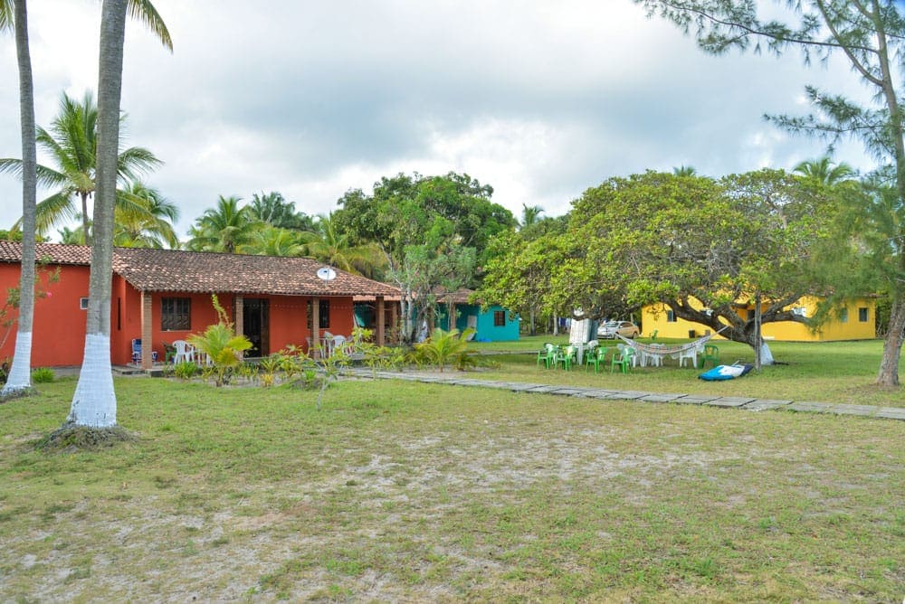 Two colorful houses are seen amidst lush greenery and palm trees. White chairs are arranged on the lawn, complemented by a large tree providing shade. A walking path made of stones guides toward the residences, creating a serene outdoor environment.
