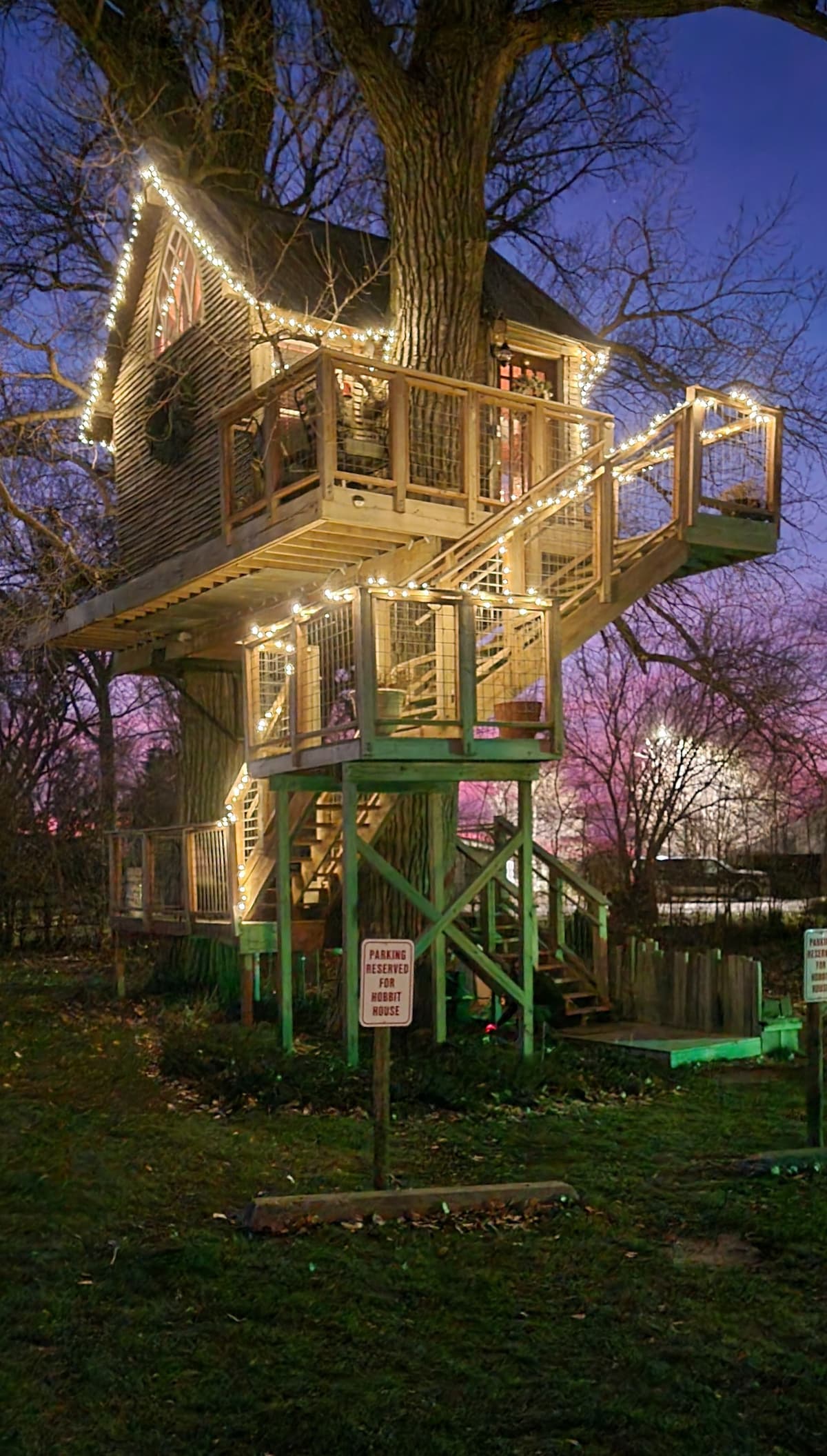 A charming treehouse is elevated among cottonwood trees, featuring wooden balconies and illuminated with warm string lights. The evening sky displays vibrant hues, enhancing the cozy ambiance. A sign is visible in the foreground, indicating property guidelines.