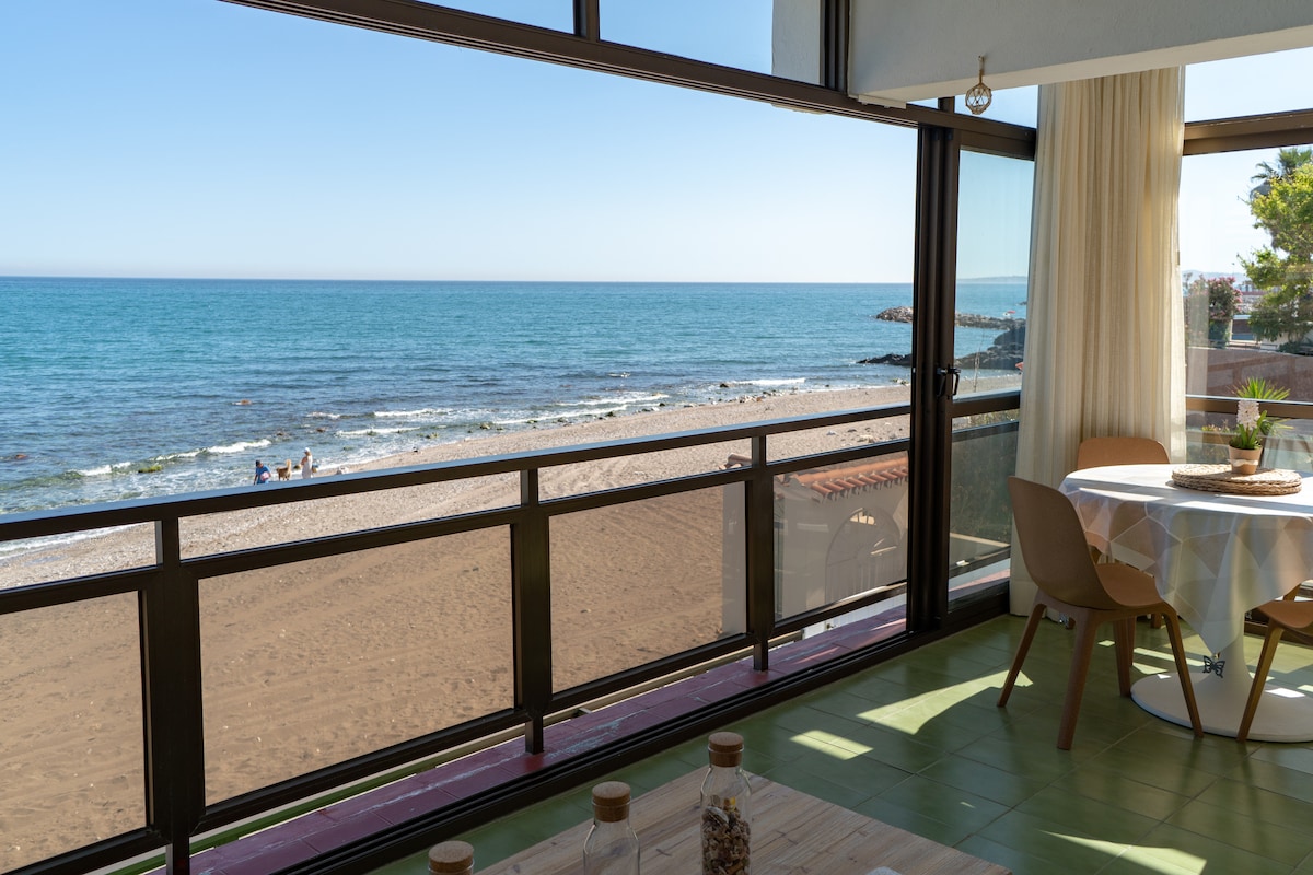 A bright dining area features large glass windows offering panoramic views of the sandy beach and blue sea. A round table is set for dining, with soft sunlight illuminating the space, creating a relaxed atmosphere. The beach is visible just beyond the window.