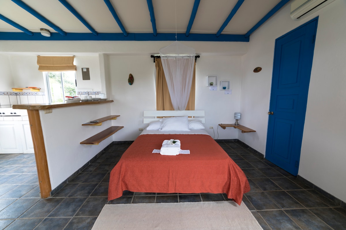 A spacious sleeping area is presented with a white bed and an orange bedspread. A small shelf is attached to the wall beside the bed, and natural light enters through a nearby window. The room features bright blue beams and a contrasting blue door.