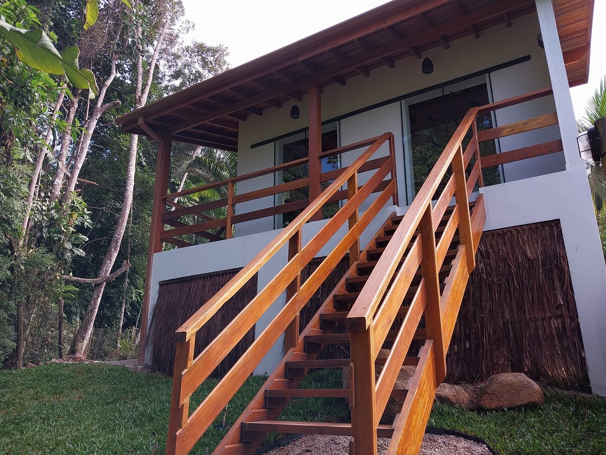 A wooden staircase leads up to an elevated loft, framed by natural landscaping. The exterior features a combination of wooden beams and white walls, with large windows allowing ample light into the space. Lush greenery surrounds the property.