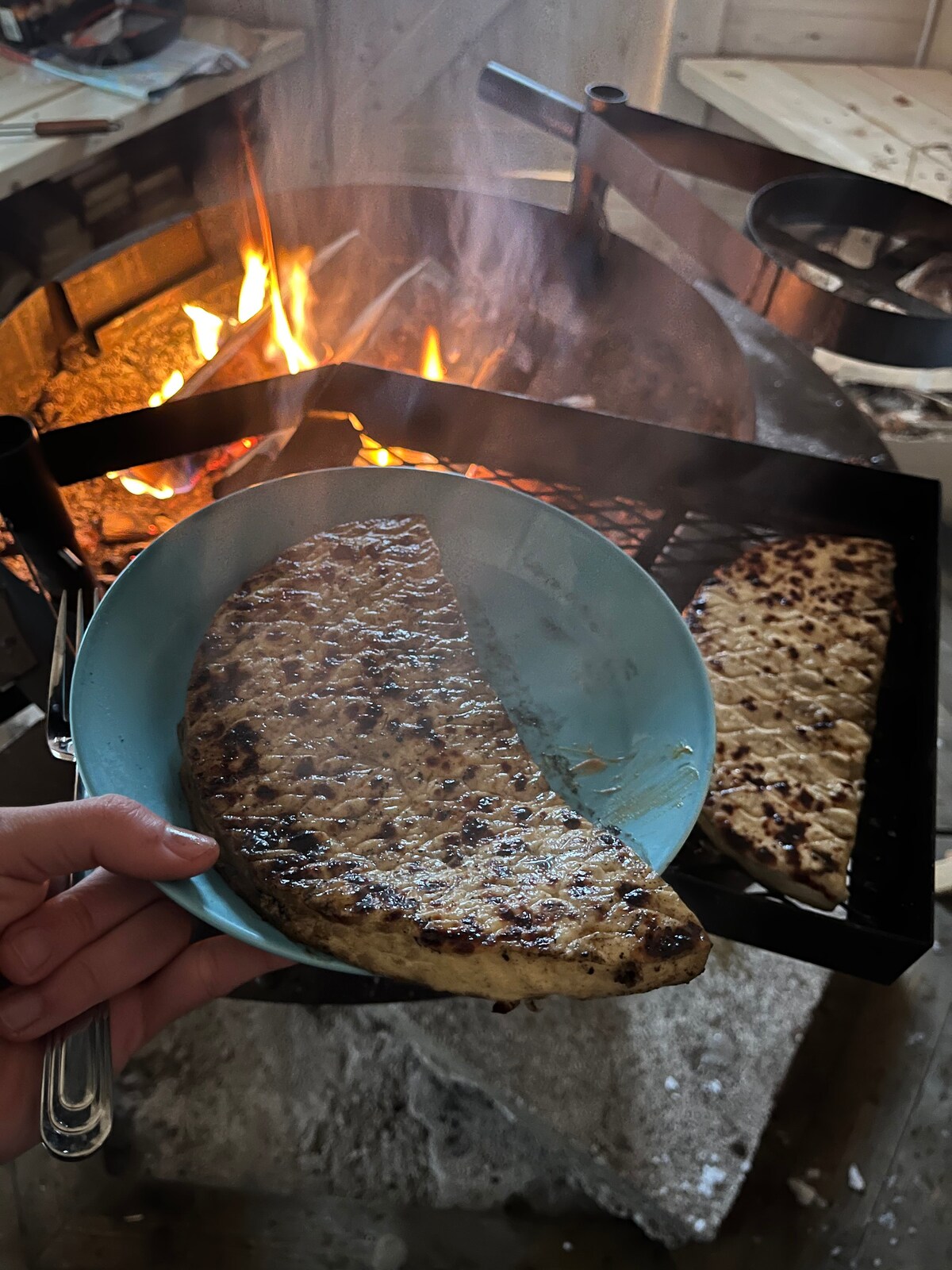A hand holds a blue plate featuring a grilled dish, showcasing a golden-brown, charred surface. In the background, a fire is visible, with flames dancing over a grill setup. The cozy indoor setting includes a rustic atmosphere with warm lighting.