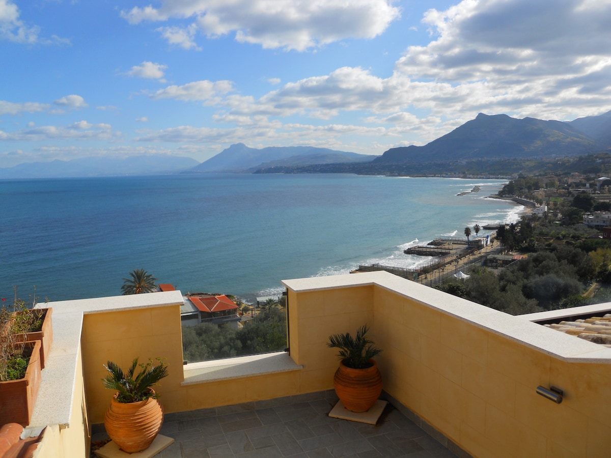 A spacious terrace features two potted plants and offers a panoramic view of the tranquil sea, with waves gently lapping at the shore. Mountains rise in the background under a partly cloudy sky, creating a serene coastal atmosphere.