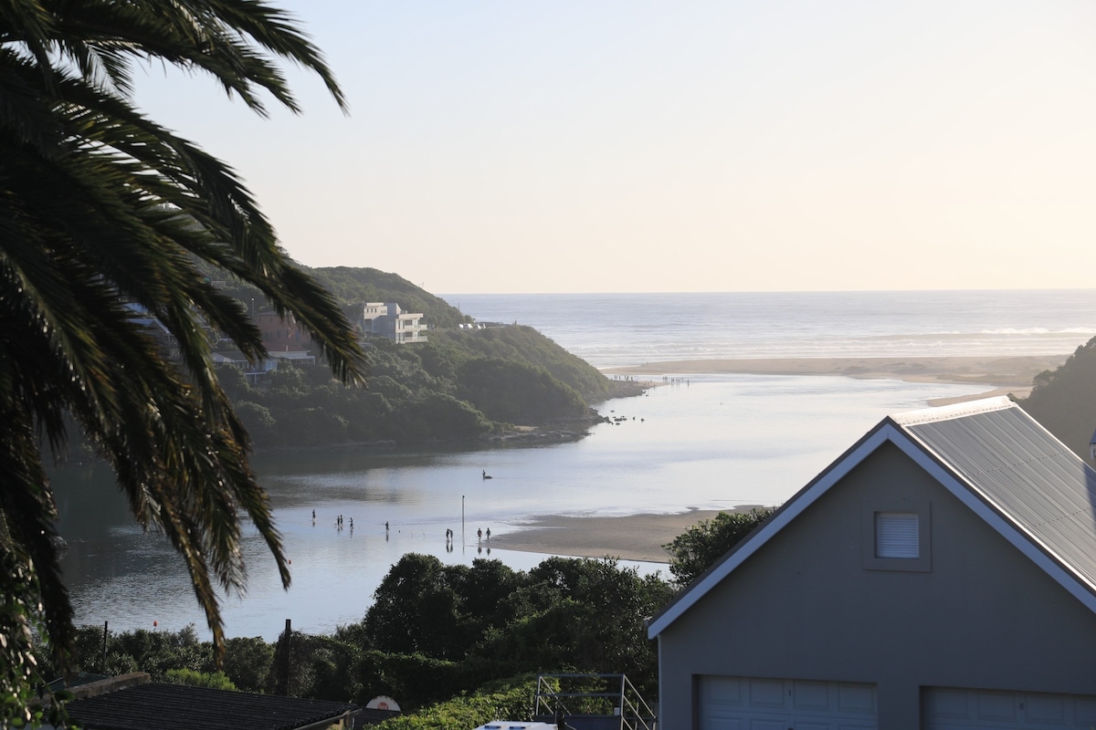A view of the lagoon meeting the sea, framed by lush greenery and palm trees. In the distance, silhouettes of people can be seen enjoying the water. The scene captures the gentle curvature of the coastline under soft morning light.