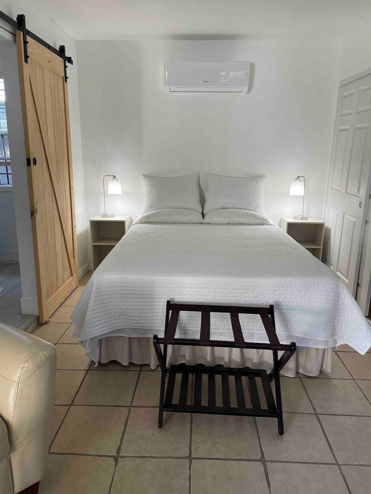 A comfortable sleeping area features a spacious bed dressed in crisp white bedding. Flanking the bed are two nightstands with lamps providing soft illumination. A stylish barn door leads to an adjacent space, while a simple folding stool is positioned at the foot of the bed.